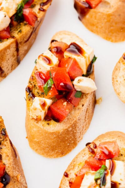 A slice of gluten-free toasted baguette topped with tomato, mozzarella, basil and balsamic glaze.