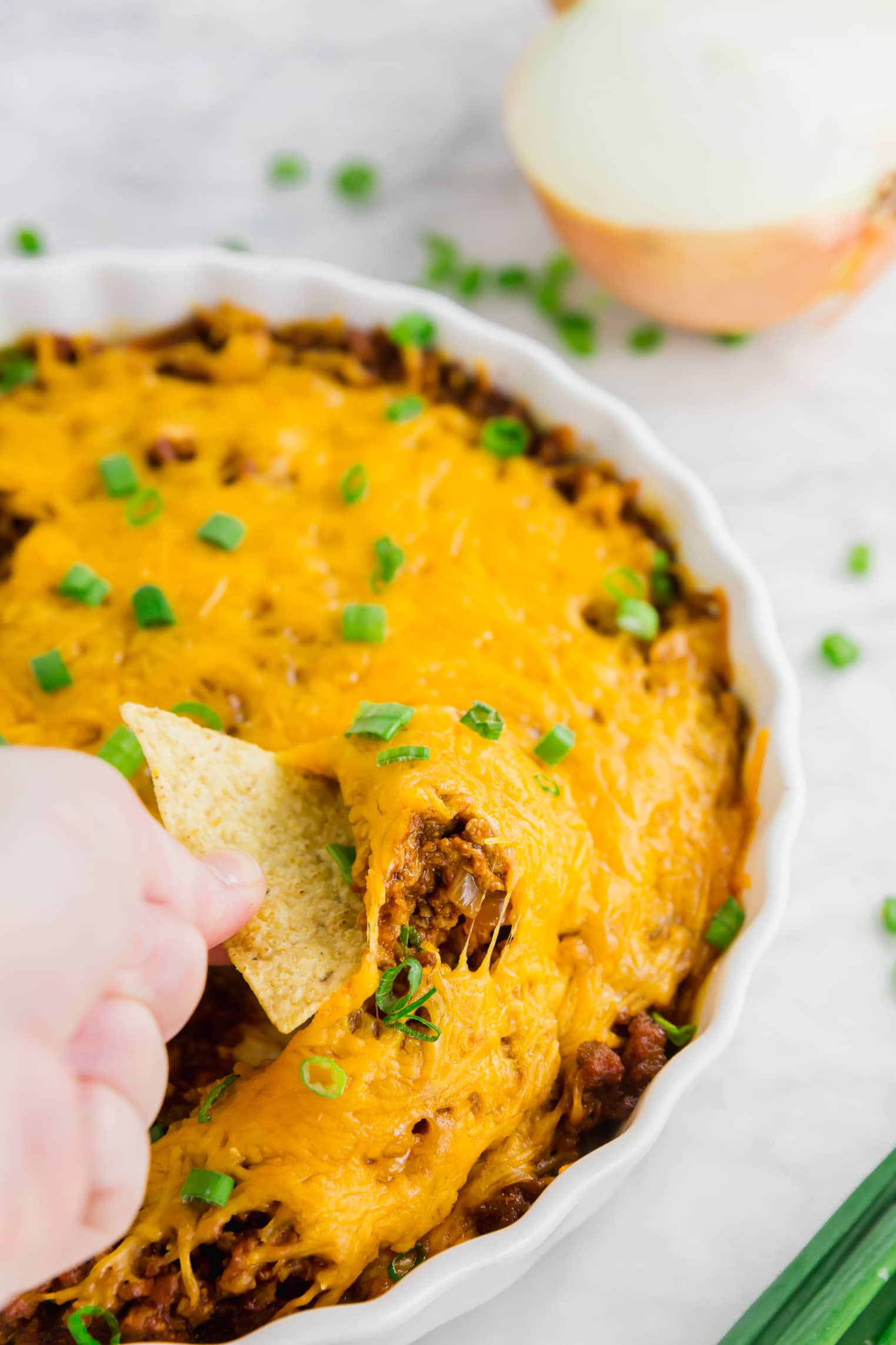 A photo of gluten-free chili cheese dip in a white tart pan with a hand dipping a tortilla into the dish. 