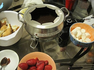A fondue pot filled with chocolate sits on a glass table, surrounded by bowls of strawberries, marshmallows, and pieces of cake, ready for dipping.