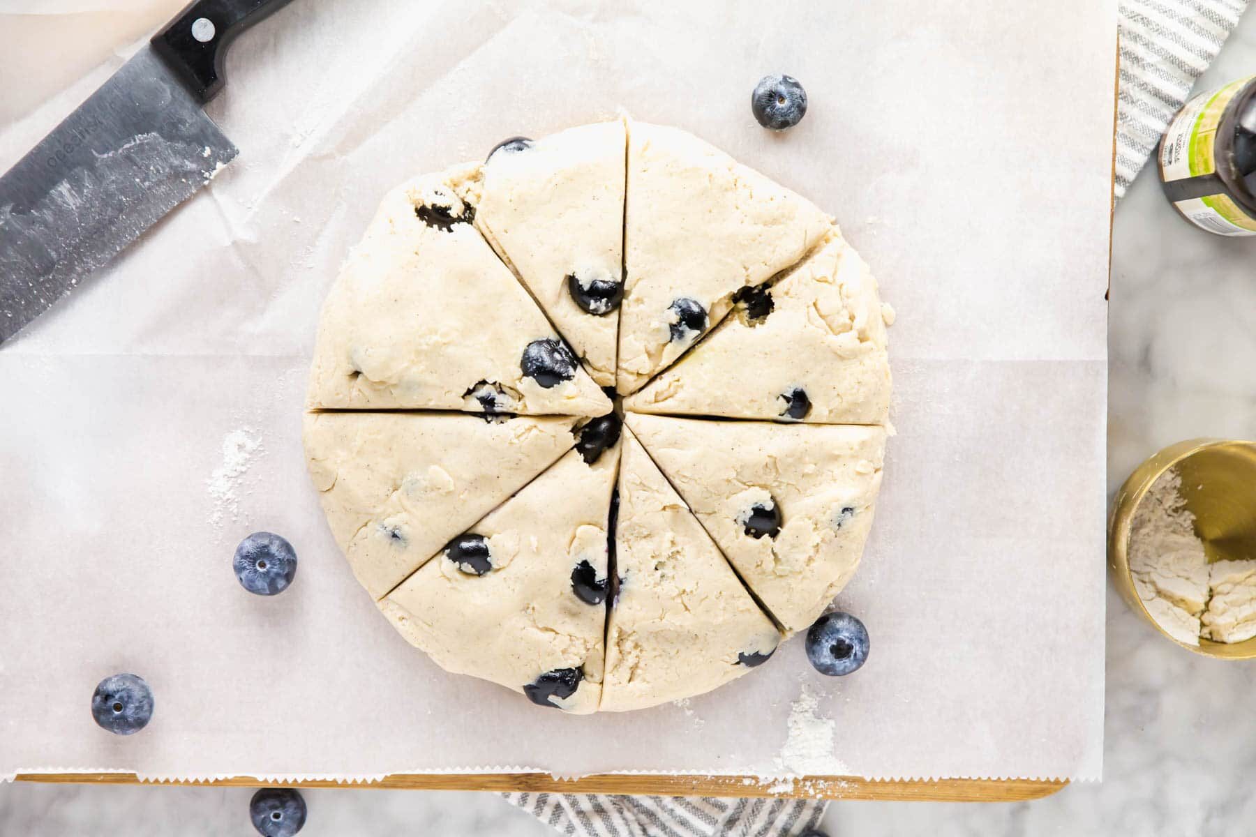 Round blueberry scone dough cut into eight wedges sits on parchment paper, with fresh blueberries scattered nearby. A large knife rests in the corner, and baking tools are partially visible around the edges.
