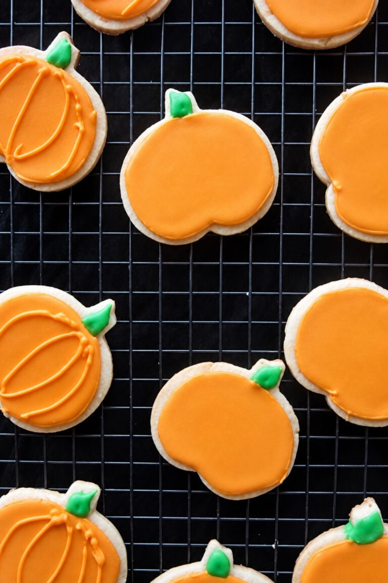 Pumpkin-shaped sugar cookies decorated with orange icing and green stems cooling on a wire rack.