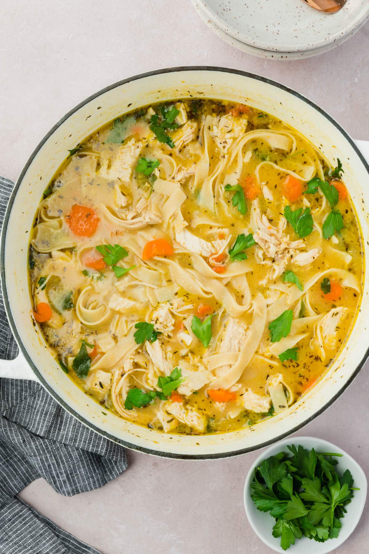 A white dutch oven with gluten free chicken noodle soup topped with fresh chopped parsley.