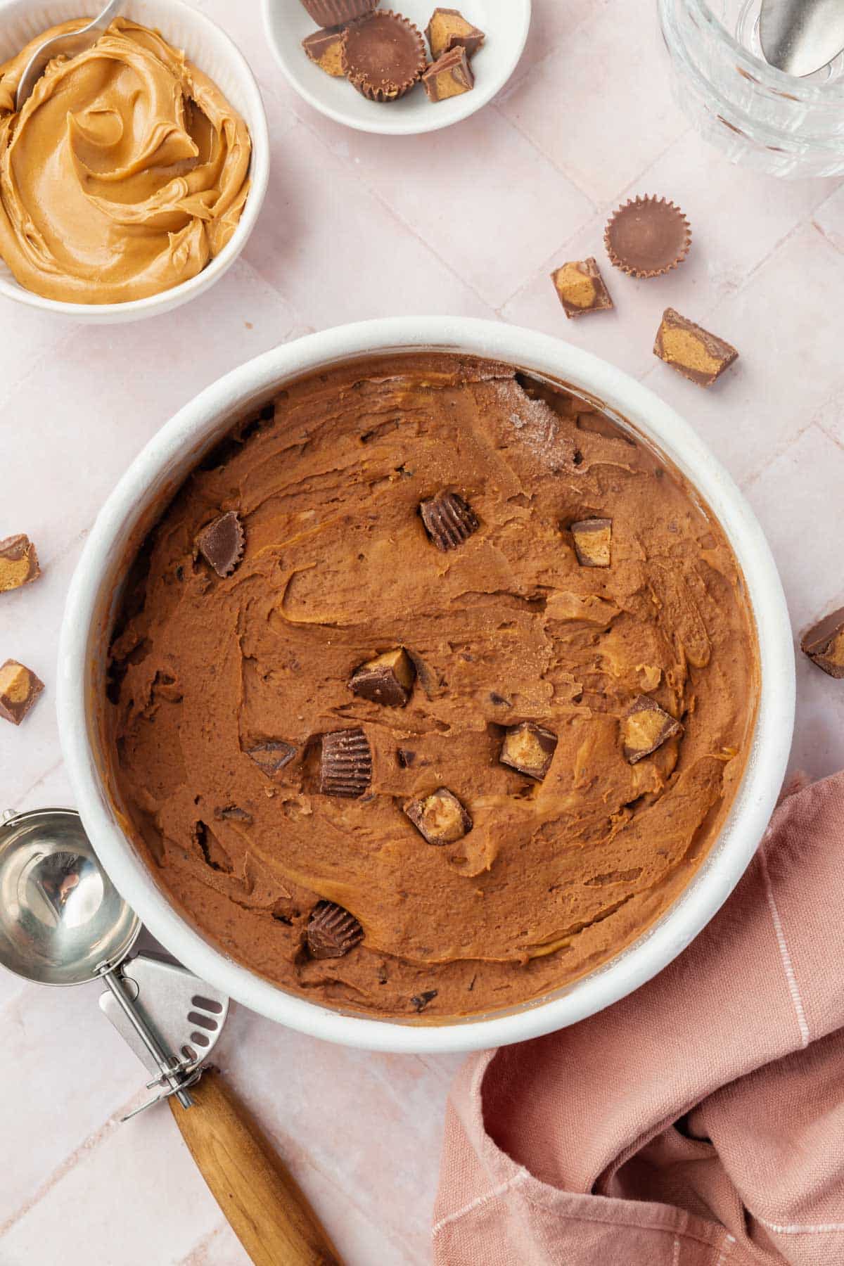 Chocolate peanut butter ice cream in a round pan surrounded by a bowl of peanut butter, a bowl of chopped peanut butter cups, a pink napkin, and a wood ice cream scooper.