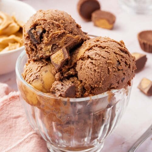 A glass bowl of scoops of chocolate peanut butter ice cream topped with peanut butter cups in front of a bowl of peanut butter and chopped up peanut butter cups.