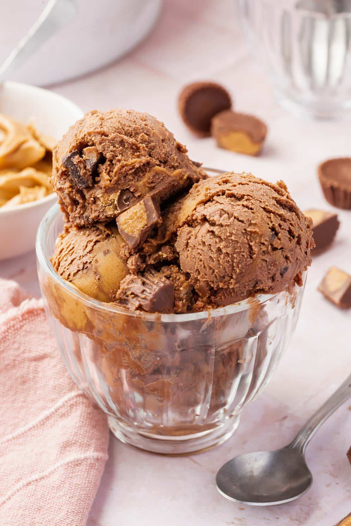 A glass bowl of scoops of chocolate peanut butter ice cream topped with peanut butter cups in front of a bowl of peanut butter and chopped up peanut butter cups. 