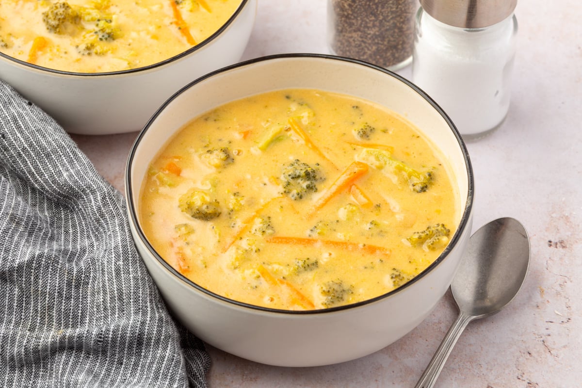 Side-angle view of a bowl of broccoli cheddar soup with carrots and broccoli, placed next to salt and pepper shakers and a striped cloth napkin.