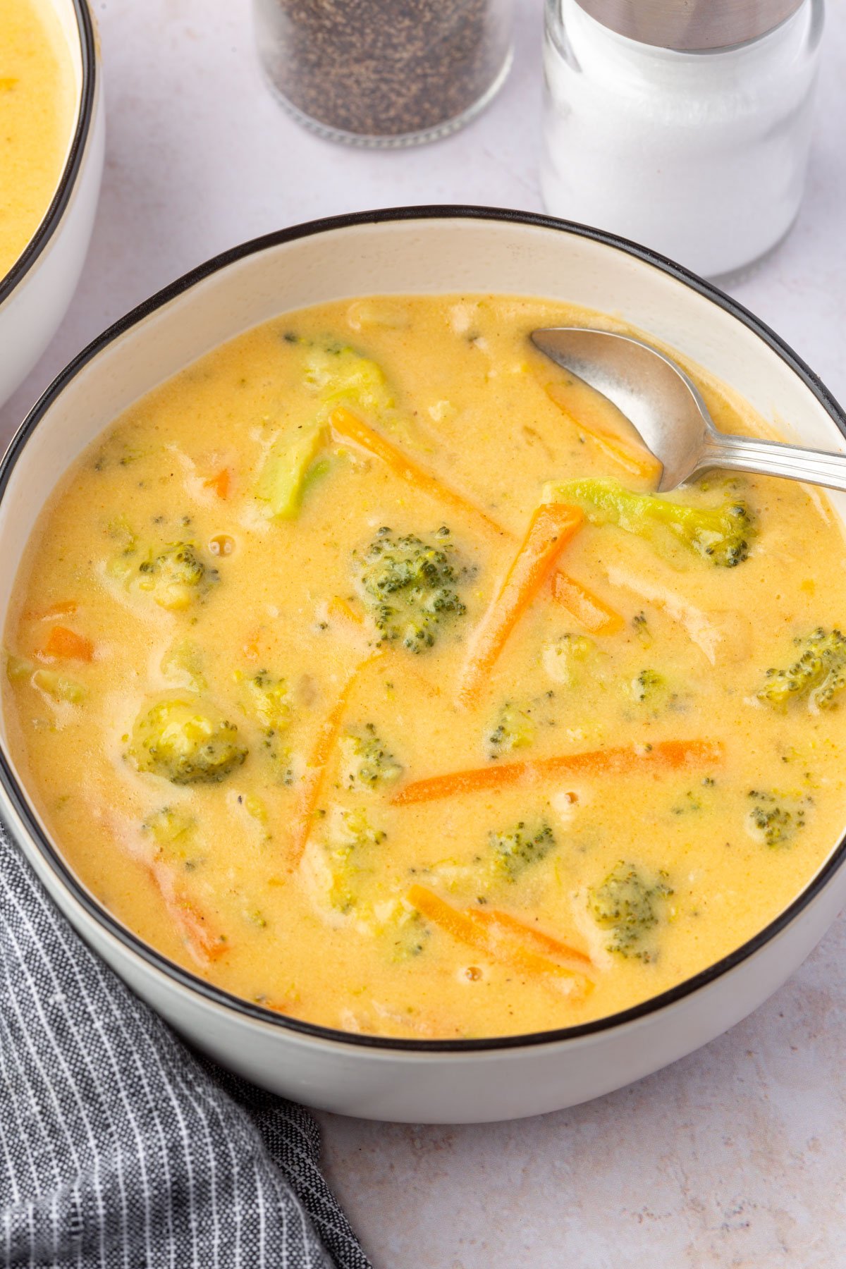 Overhead shot of a bowl of creamy broccoli cheddar soup with a spoon resting inside and salt and pepper shakers in the background.