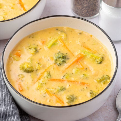 Overhead shot showing two bowls of broccoli cheddar soup on a white countertop, next to a spoon and black striped linen napkin.
