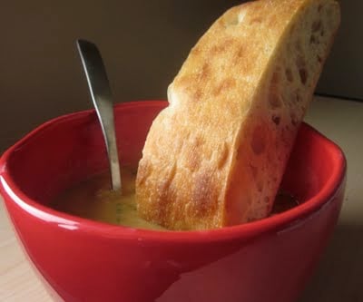 Broccoli cheddar soup in a red bowl with a spoon and gluten-free ciabatta bread.