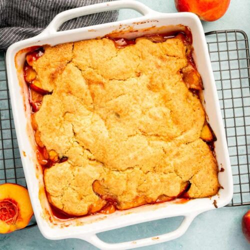 An overhead view of a square baking dish of gf peach cobbler with fresh peach halves around the dish.