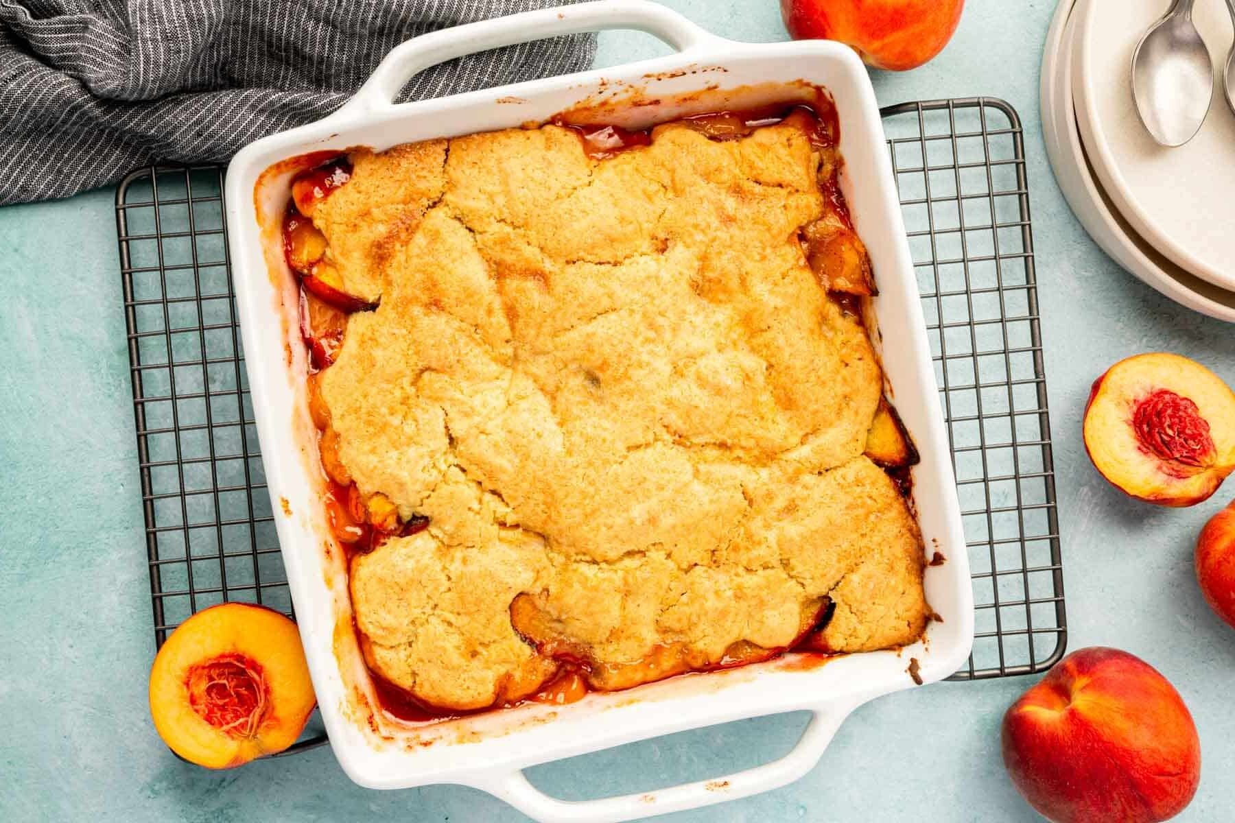 An overhead view of a square baking dish of gf peach cobbler with fresh peach halves around the dish.