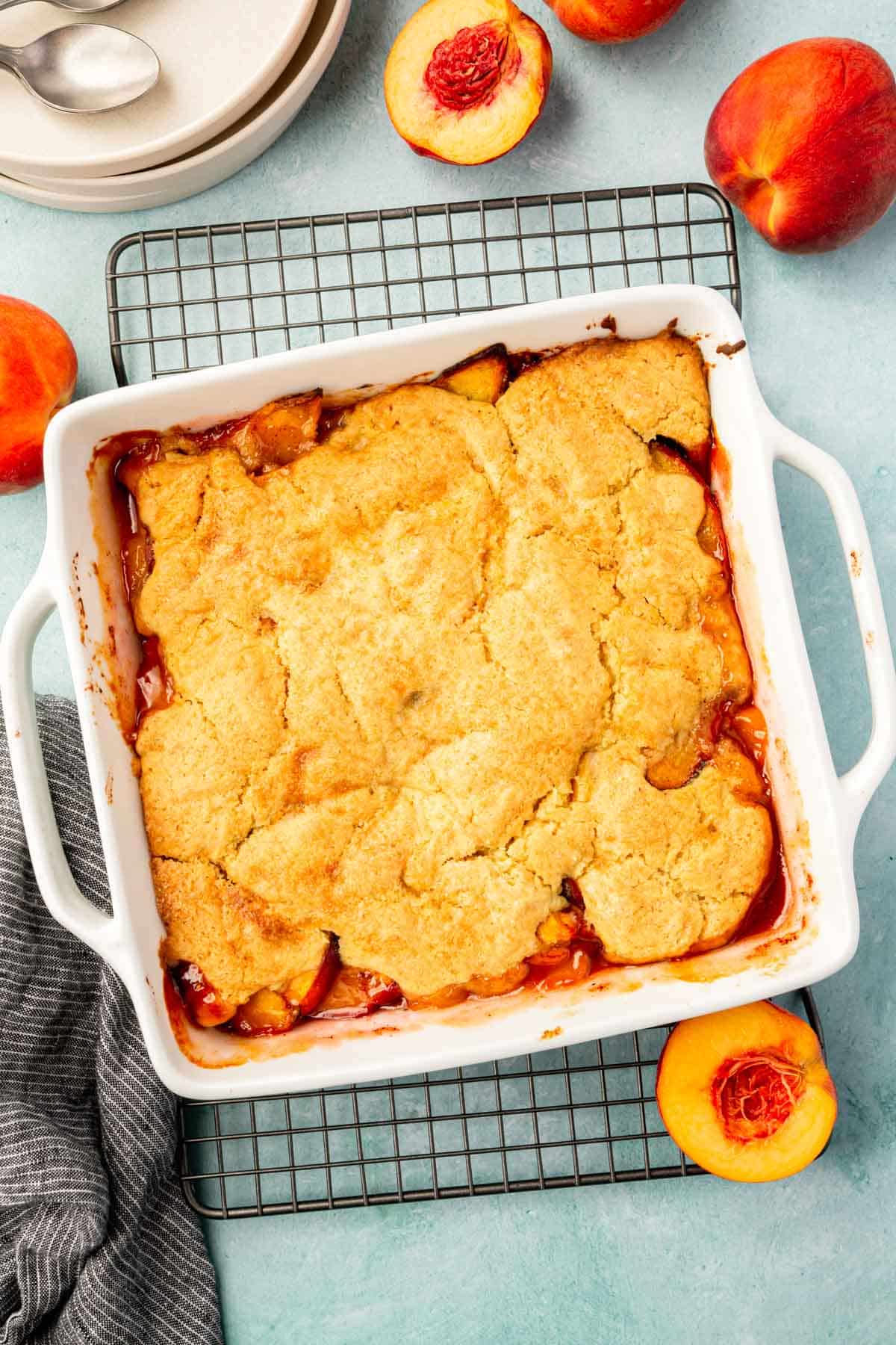 An overhead view of a square baking dish of gf peach cobbler with fresh peach halves around the dish.