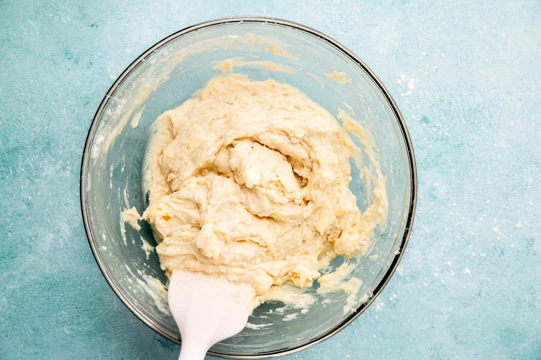 A glass mixing bowl with cobbler biscuit batter and a spatula.
