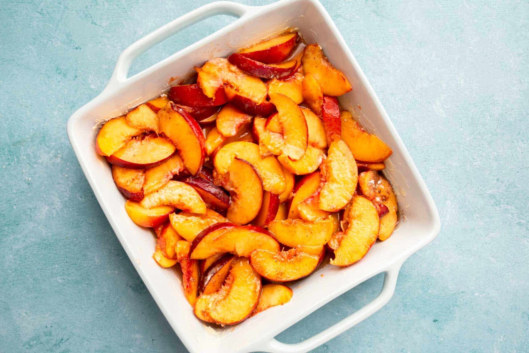 A square baking dish filled with sliced peach filling for cobbler.