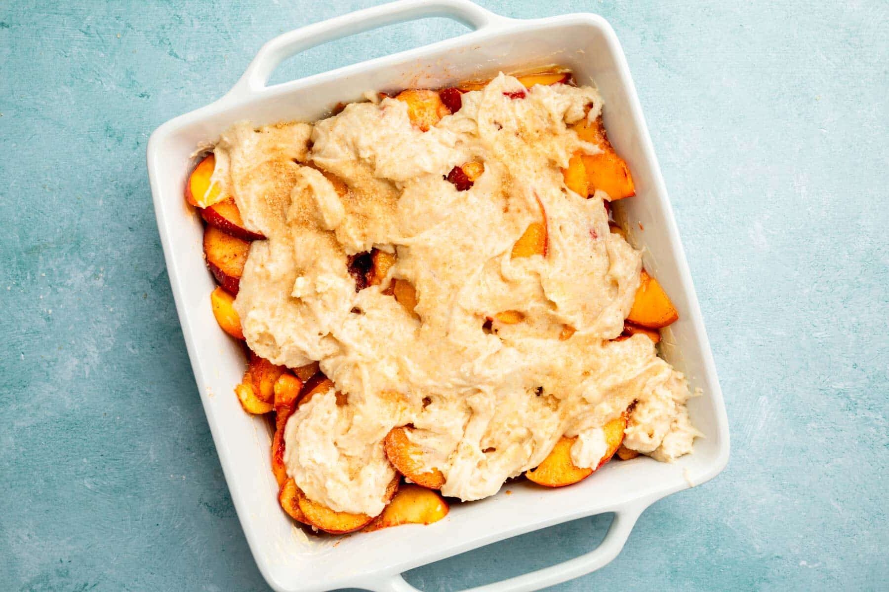 A square baking pan with peach filling and biscuit batter on top before baking.