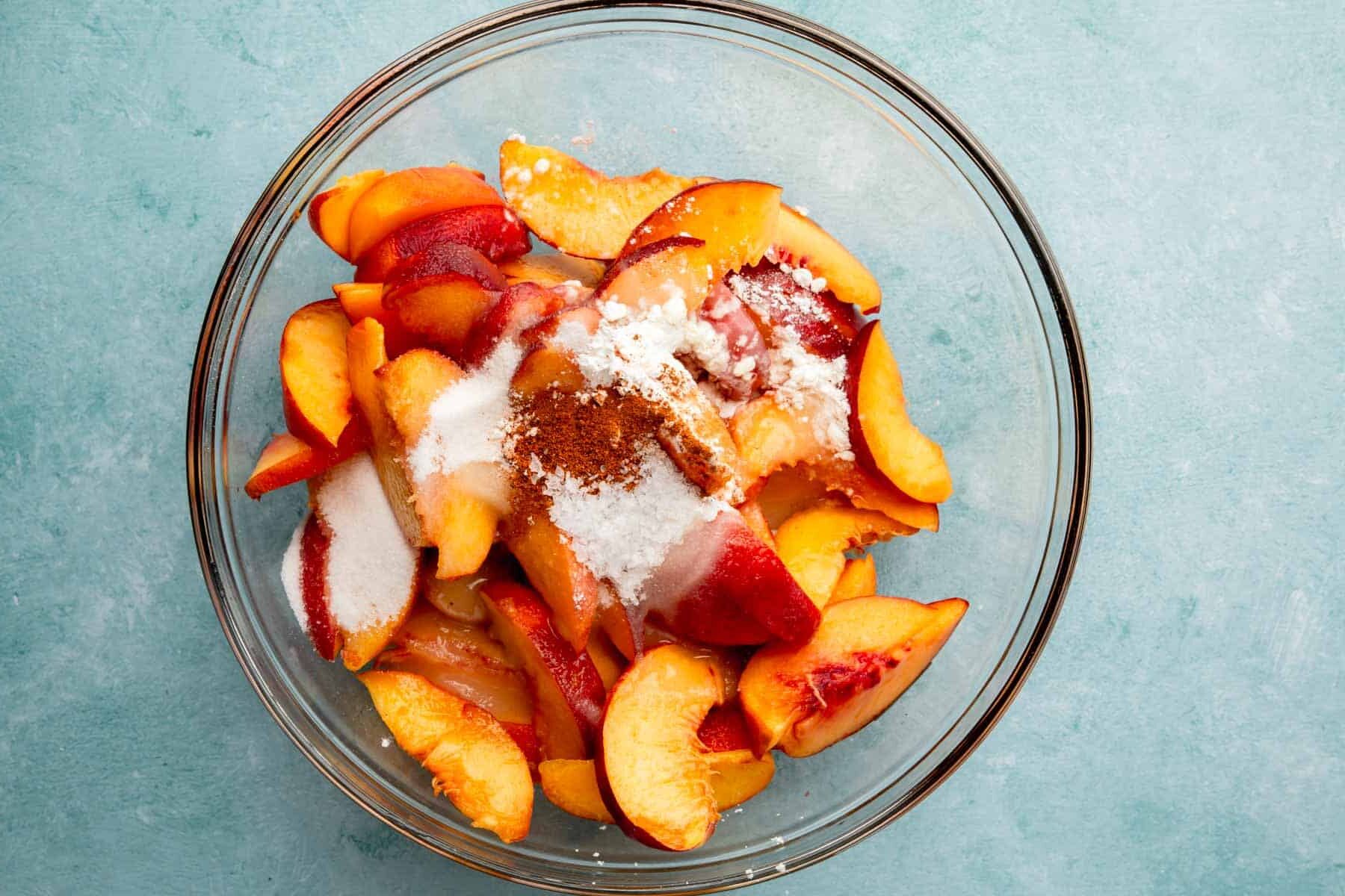 A glass mixing bowl with sliced peaches, cornstarch, lemon juice, cinnamon, nutmeg, and salt before mixing.