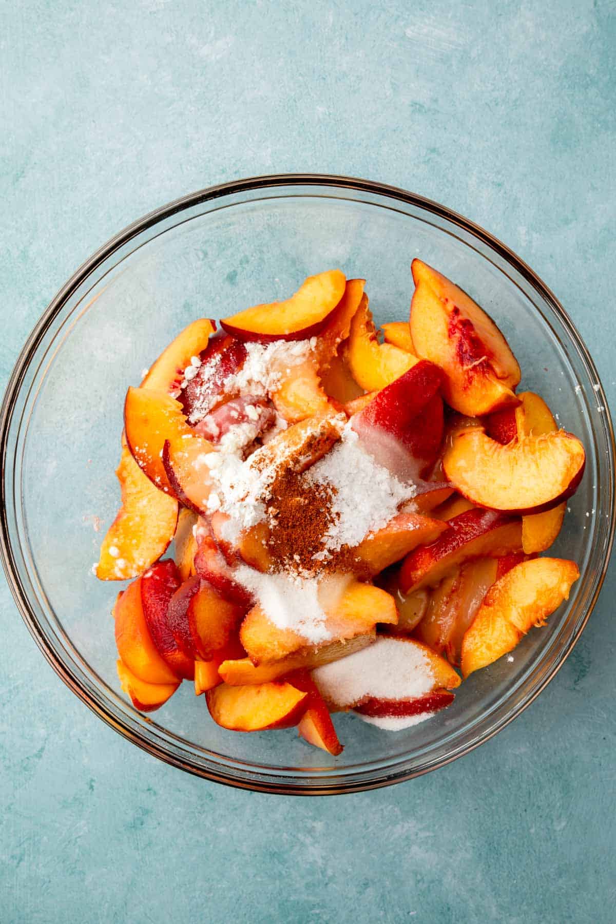 A glass mixing bowl with sliced peaches, cornstarch, lemon juice, cinnamon, nutmeg, and salt before mixing.