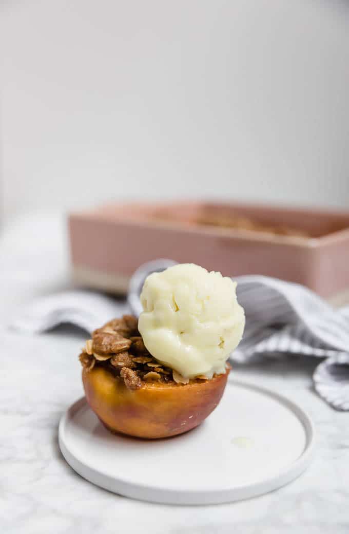 Straight on view of a peach on a plate stuffed with streusel topping and a scoop of ice cream with a pink baking dish in the background. 