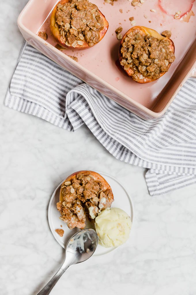 An aerial view of a baking dish with halved peaches with gluten-free streusel topping and a plate with a peach with streusel topping and a scoop of ice cream with a spoon ready to dig in. 