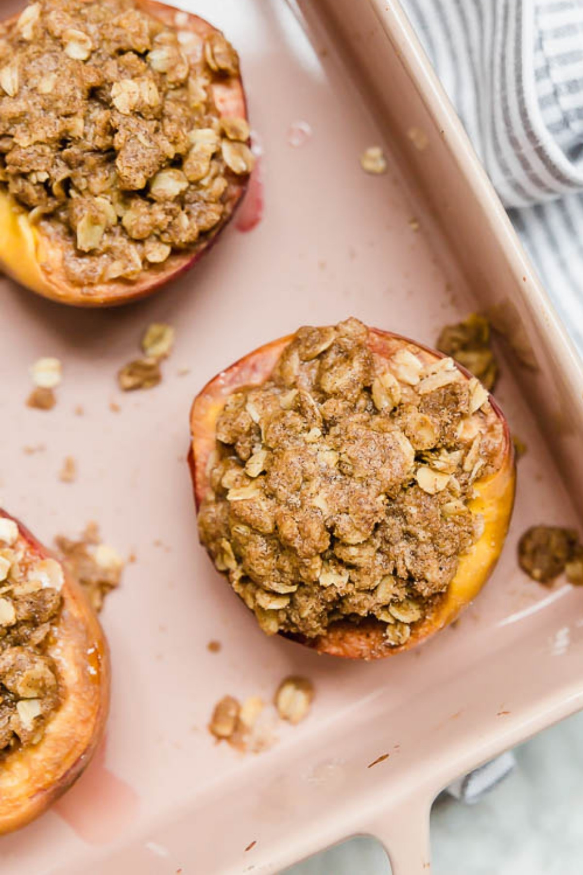 A close-up of Baked Peaches with Gluten-Free Streusel in a light pink dish. The halved peaches are topped with oat crumble, while a striped kitchen towel is partially visible in the corner.