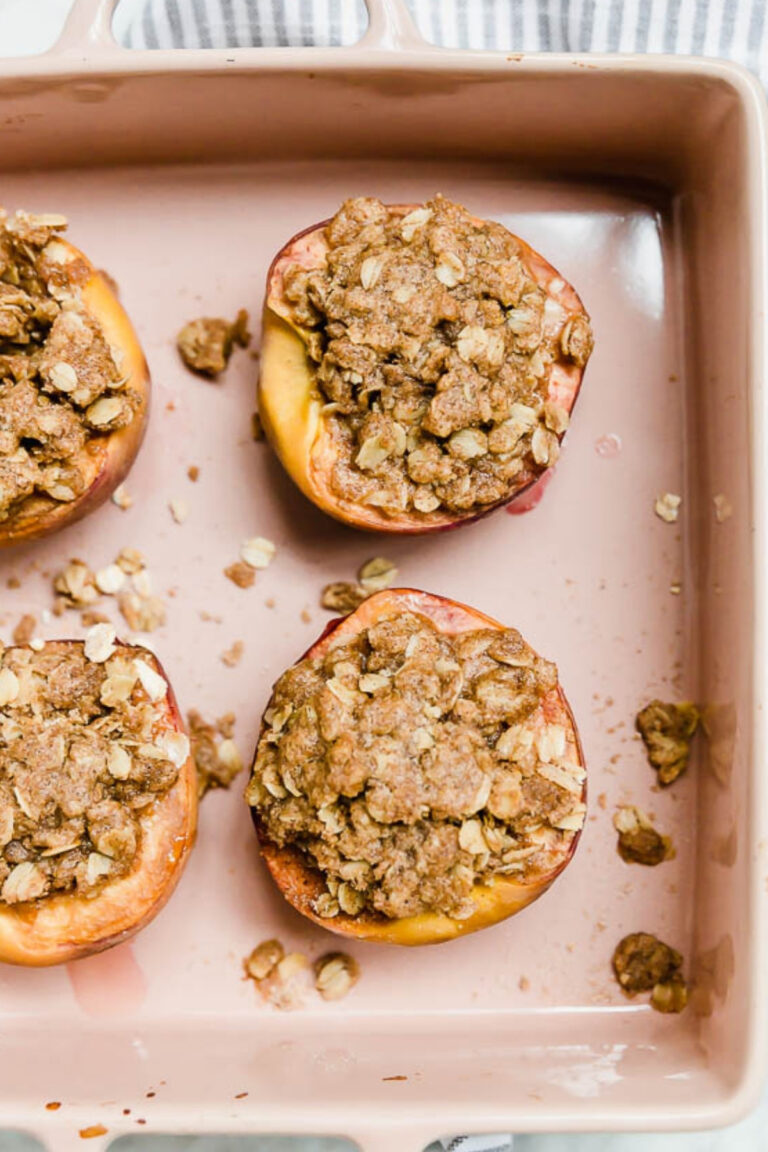 Four Baked Peaches with Gluten-Free Streusel are topped with crumbly oats and arranged in a pink baking dish, with a few golden crumbs scattered around the peaches.