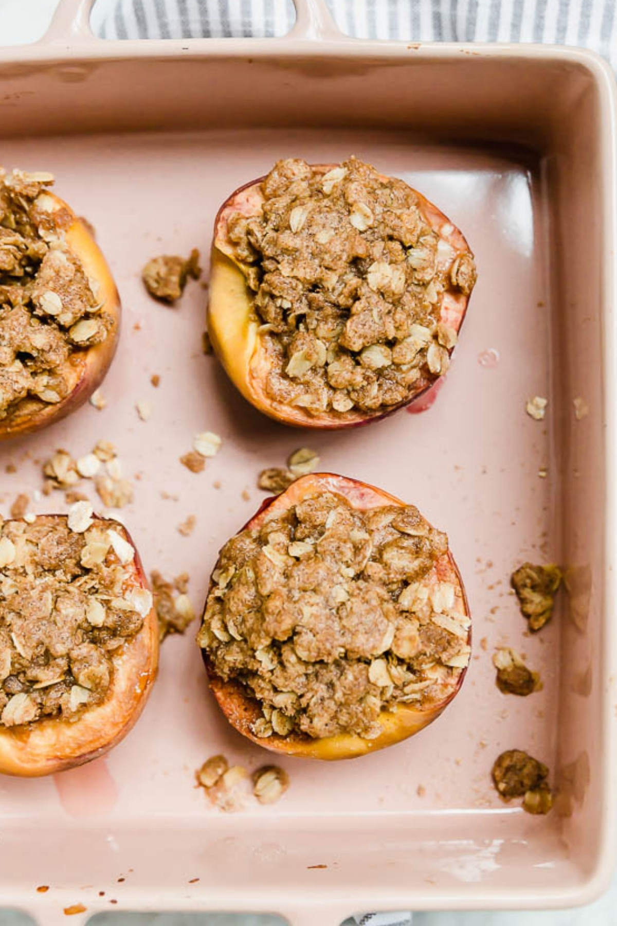 Four Baked Peaches with Gluten-Free Streusel are topped with crumbly oats and arranged in a pink baking dish, with a few golden crumbs scattered around the peaches.