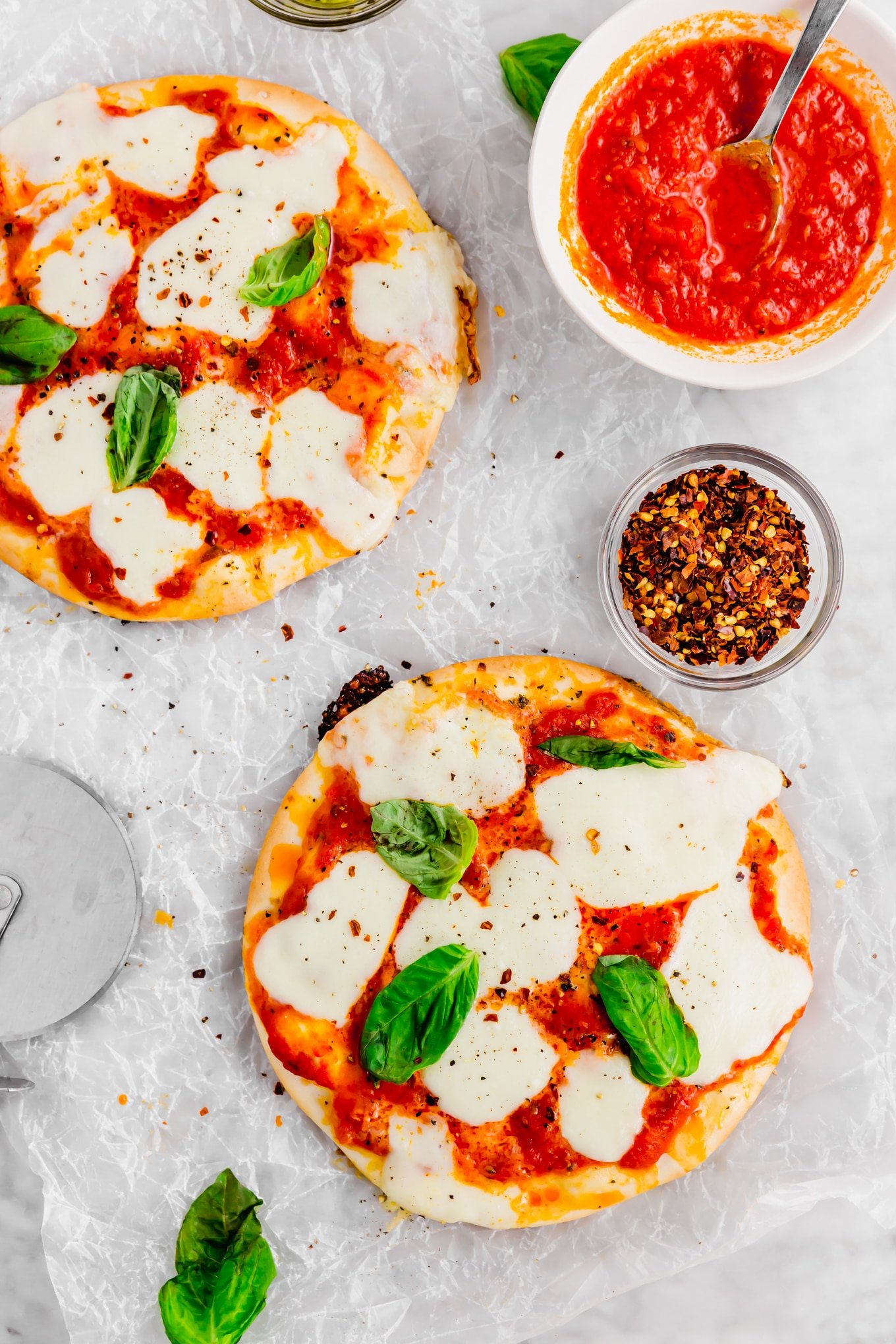 An overhead photo of two pita pizzas topped with marinara sauce, fresh mozzarella, and basil with a bowl of red pepper flakes and marinara sauce. 