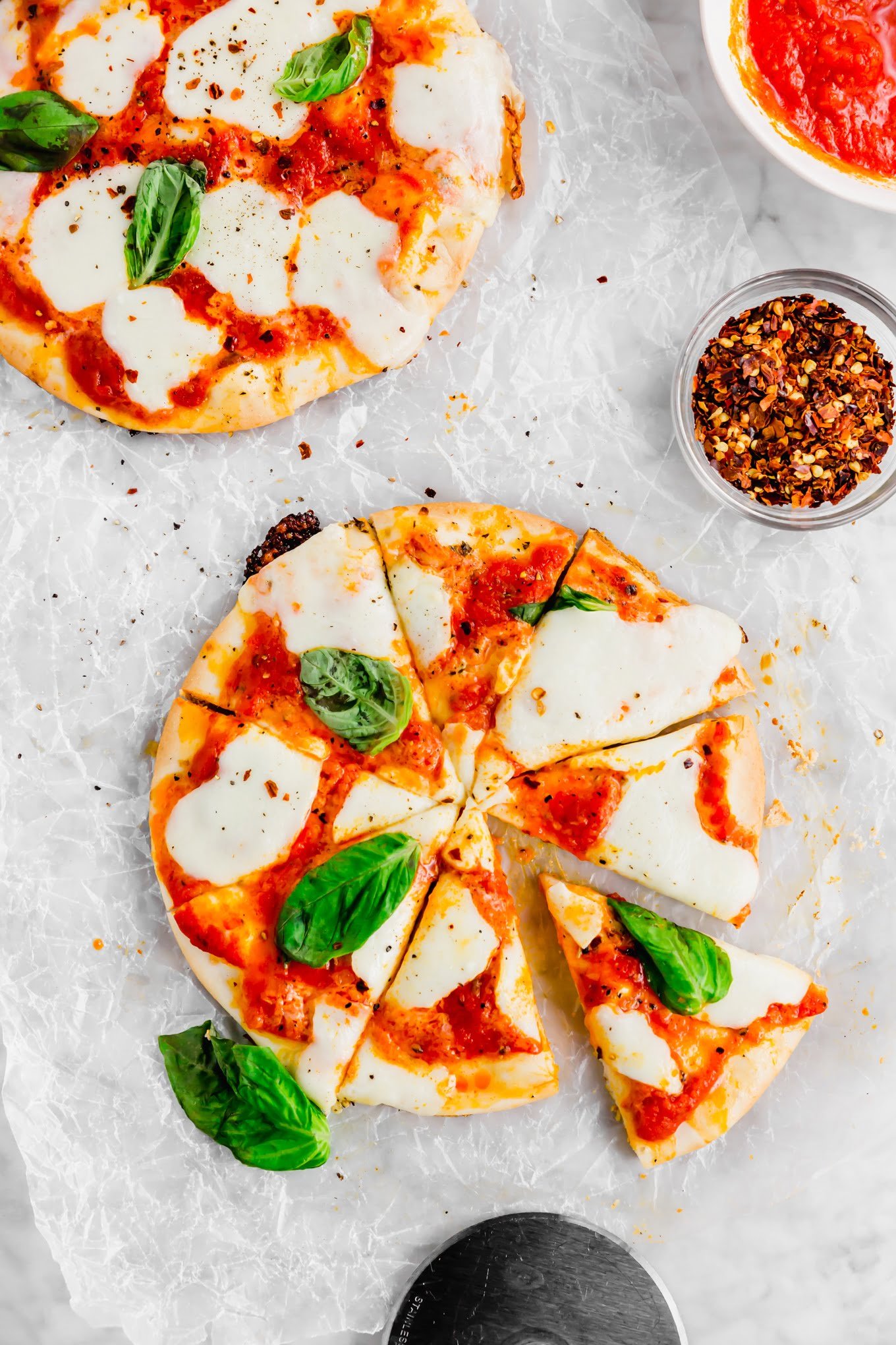 An aerial photo of two gluten-free pita pizzas topped with fresh mozzarella and basil sliced into 8 pieces with a small bowl of crushed red pepper flakes. 