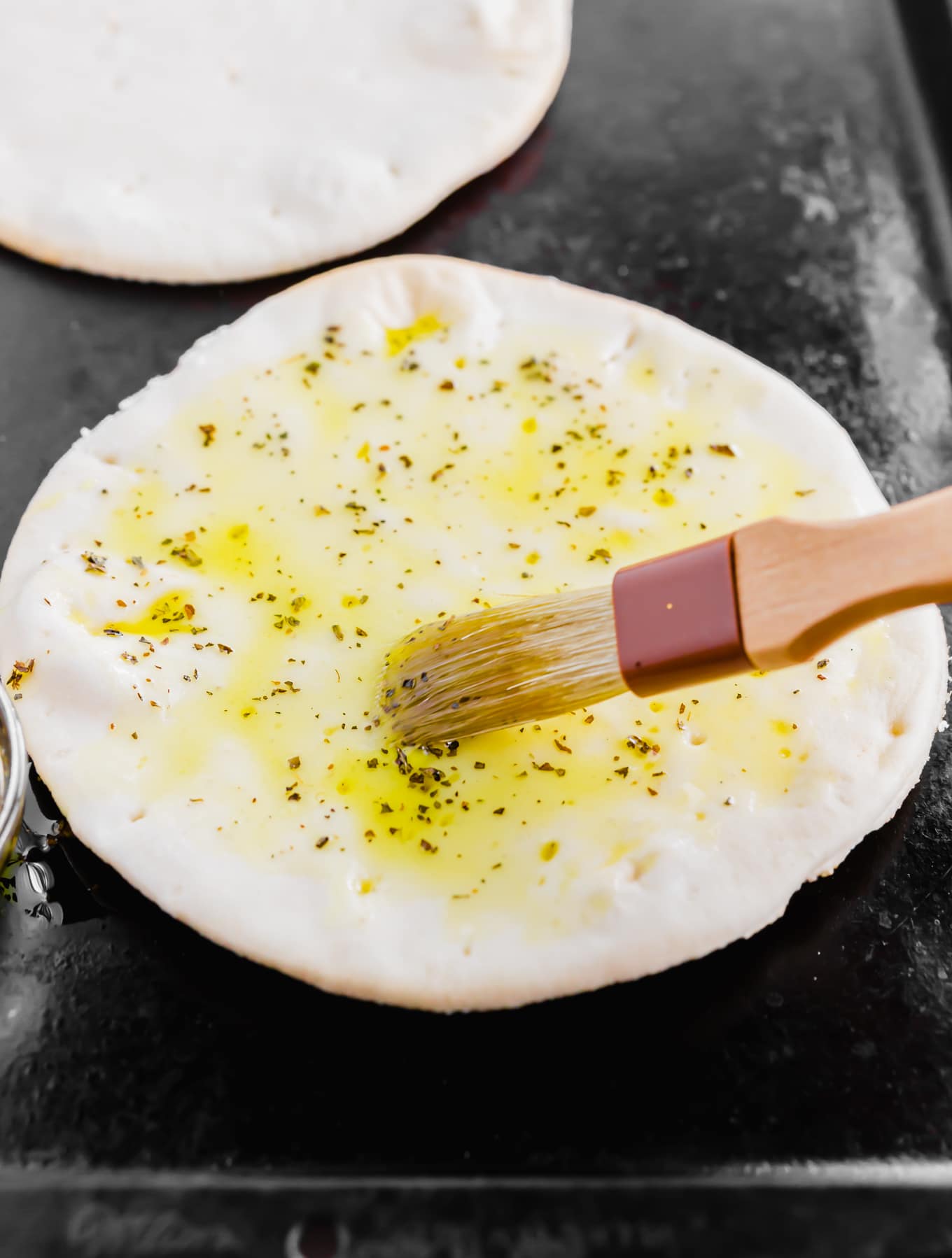 An aerial photo showing a pastry brush spreading an olive oil and herb mixture on gluten-free pita bread. 