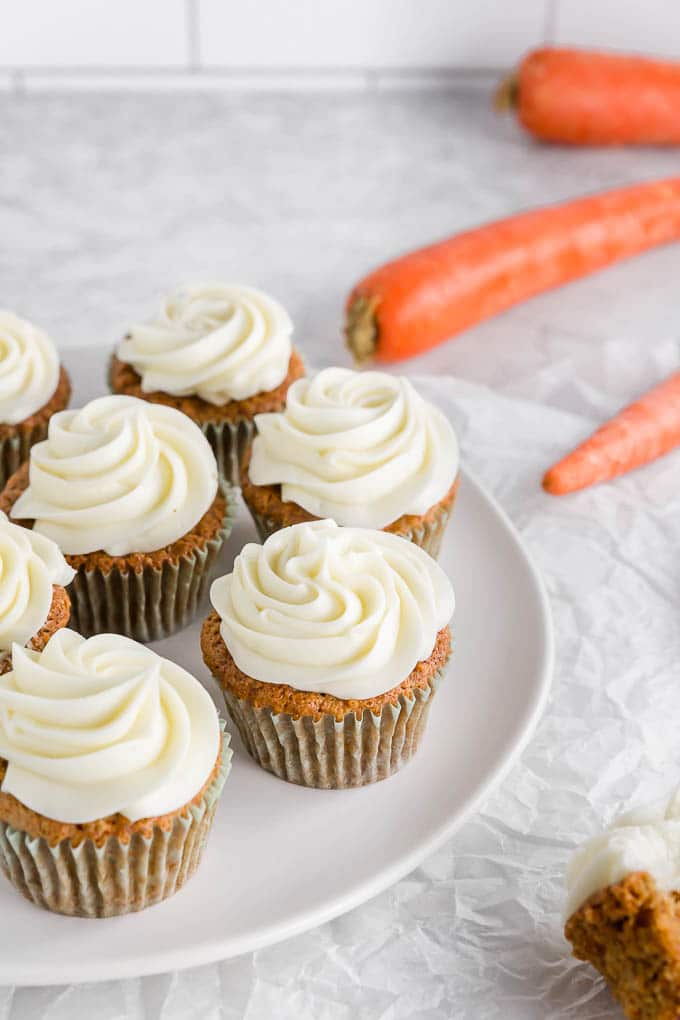A platter with a dozen gluten free carrot cake cupcakes with cream cheese frosting with large carrots in the background.