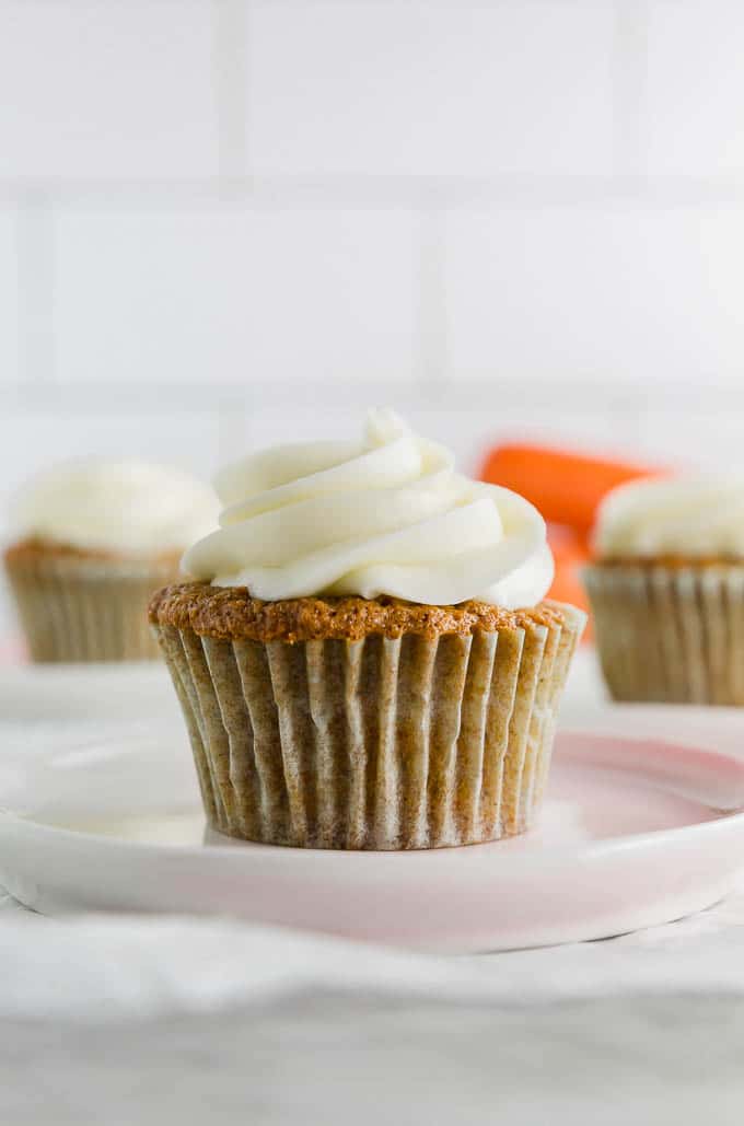 A photo of a gluten free carrot cake cupcake with cream cheese frosting on a plate with carrots in the background.