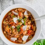 A photo of italian sausage white bean stew in a bowl topped with parmesan cheese, fresh basil and a spoon.