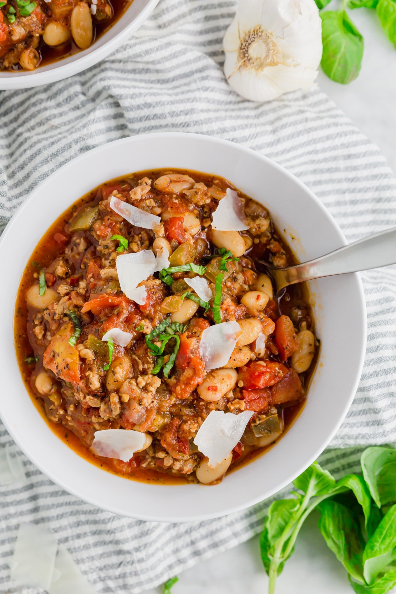 A photo of italian sausage white bean stew in a bowl topped with parmesan cheese, fresh basil and a spoon.