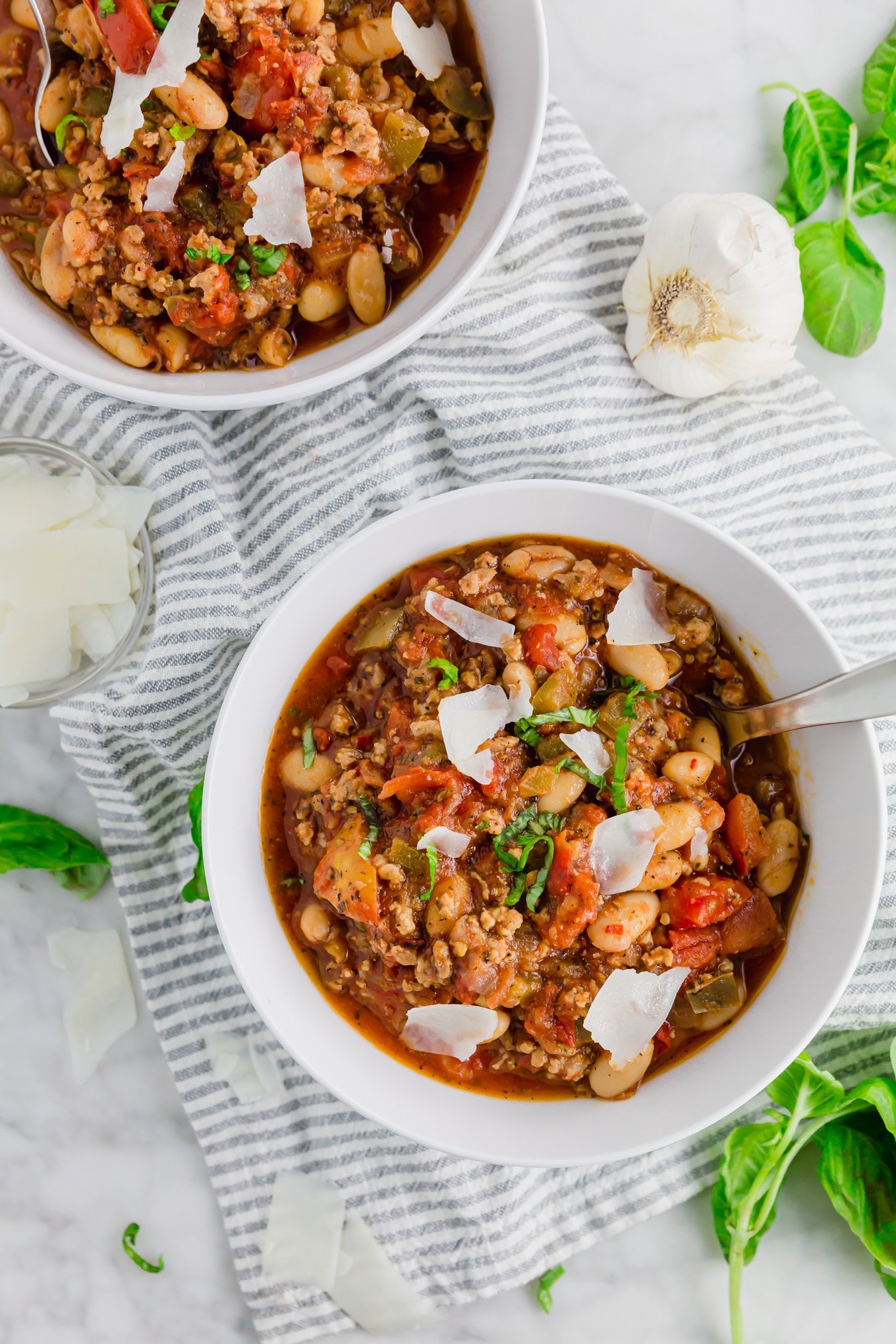 A photo of two bowls of white bean italian sausage stew topped with parmesan and fresh basil with some garlic. 