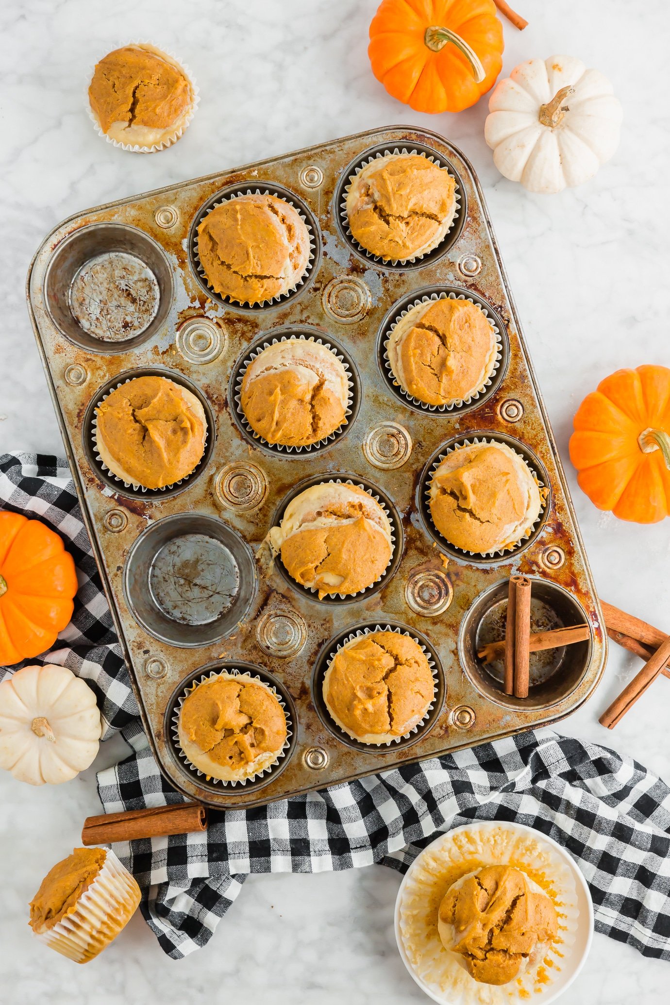 A photo of a muffin tin filled with gluten-free pumpkin cream cheese muffins and cinnamon sticks and mini pumpkins. 