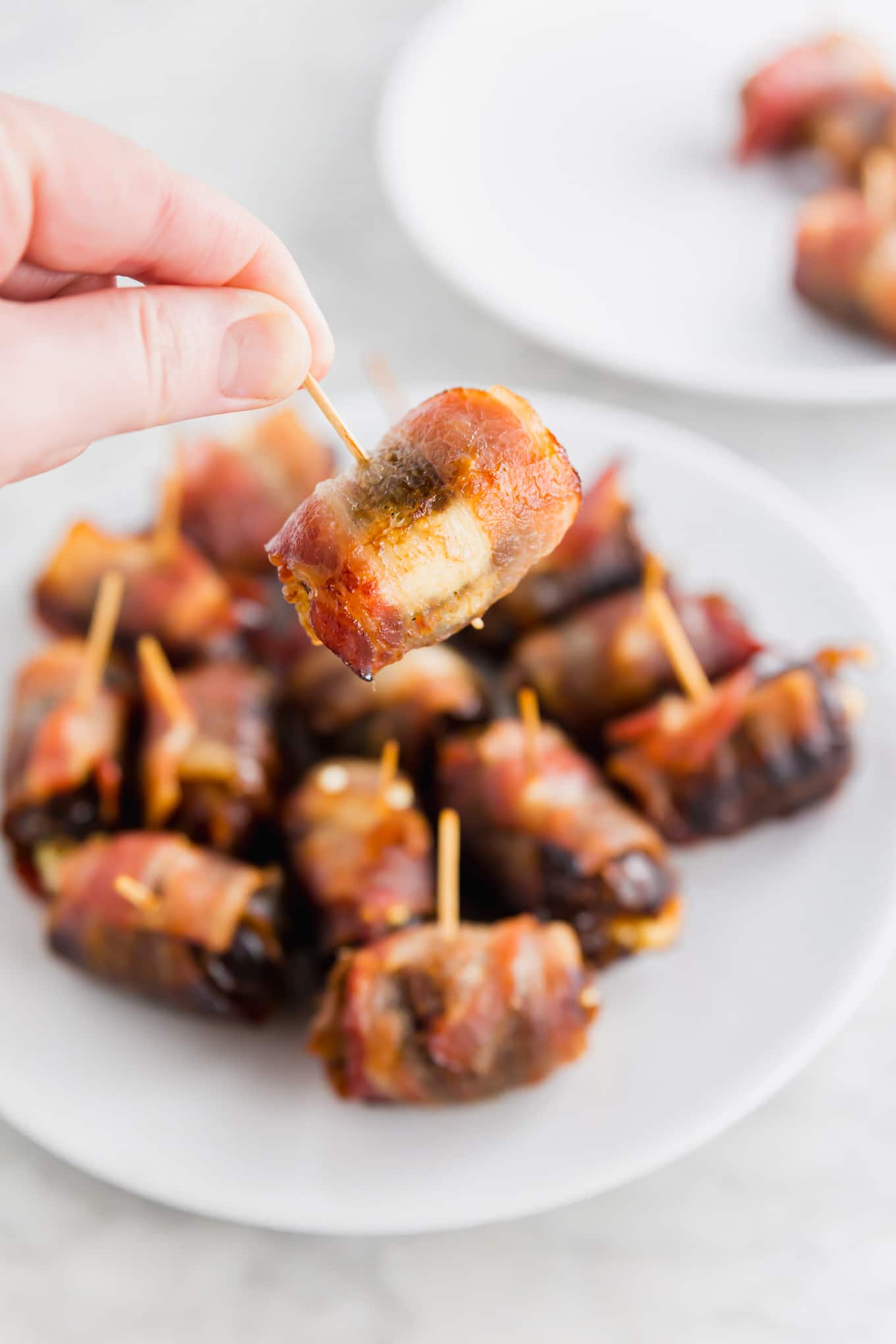 A photo of a hand holding a bacon wrapped date over a platter of goat cheese stuffed dates. 