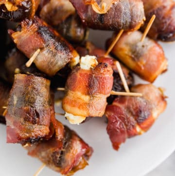 A photo of 3-ingredient bacon wrapped dates with goat cheese with toothpicks inserted in the center on a platter.