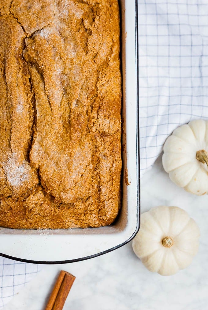 A white loaf pan with gluten-free dairy-free pumpkin bread and white mini pumpkins and a cinnamon stick. 
