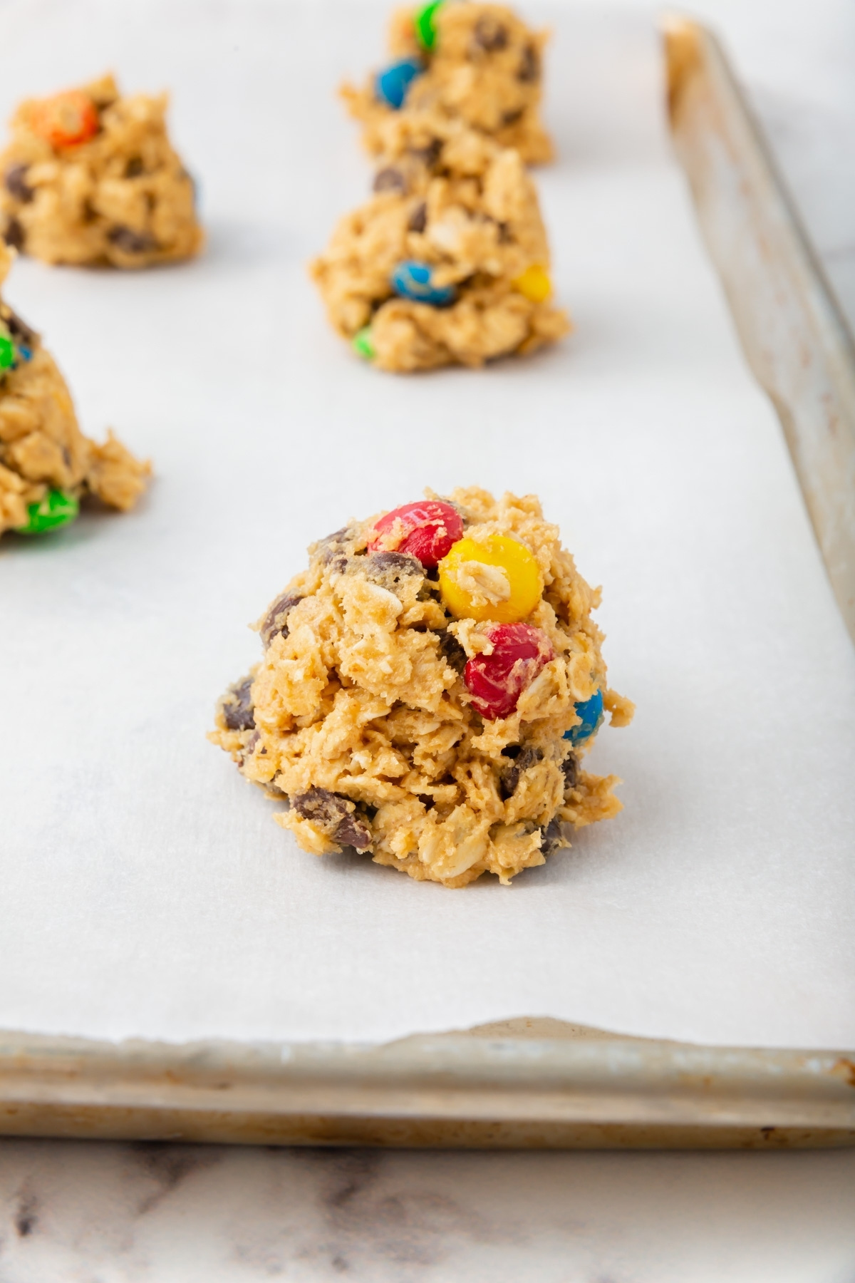 A scoop of unbaked gluten-free monster cookie dough with colorful candy-coated chocolate pieces sits on a parchment-lined baking sheet, with more dough scoops in the background.