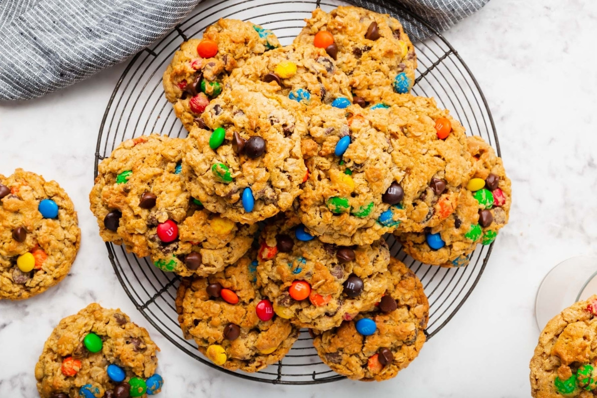A pile of colorful gluten-free monster cookies with chocolate chips and candy-coated chocolates sits on a round cooling rack, with a few cookies scattered on a white marble surface nearby.