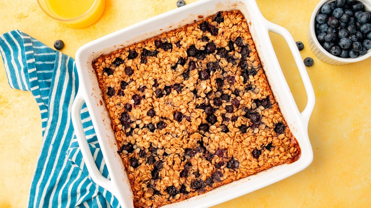 An overhead view of a baking dish with blueberry baked oatmeal in it with two glasses of orange juice and a bowl of blueberries on the side.
