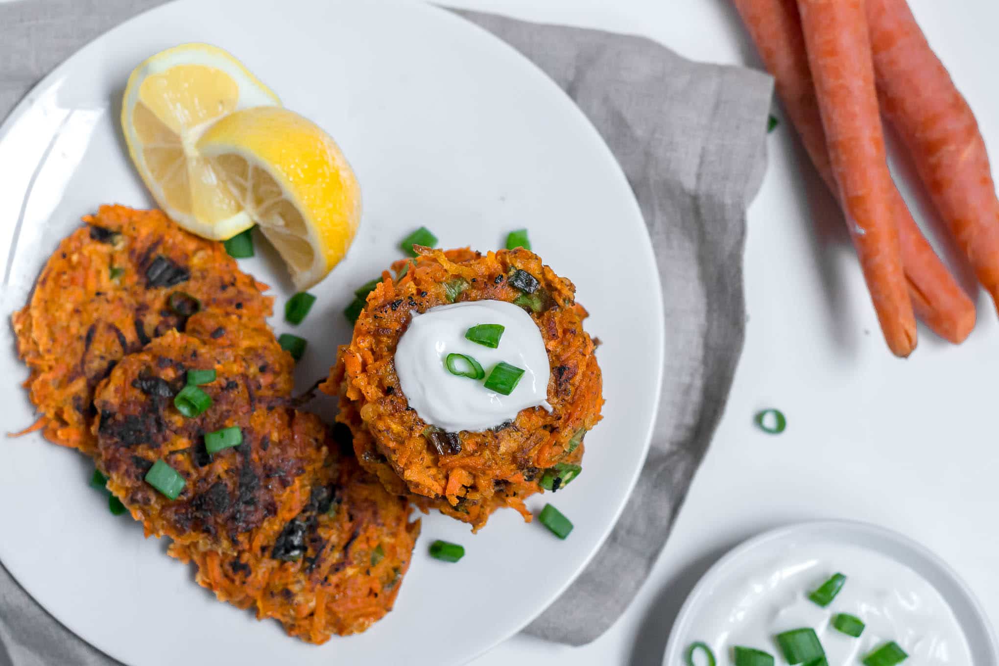 An aerial view of a white plate filled with gluten-free vegan carrot fritters topped with yogurt and green onions and lemon wedges. A gray napkin is on a white table with raw carrots int he corned and a small bowl of greek yogurt and sliced green onion. 