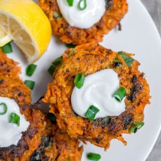 An aerial view of gluten-free carrot fritters on a white plate topped with greek yogurt, and green onions with a lemon wedge.