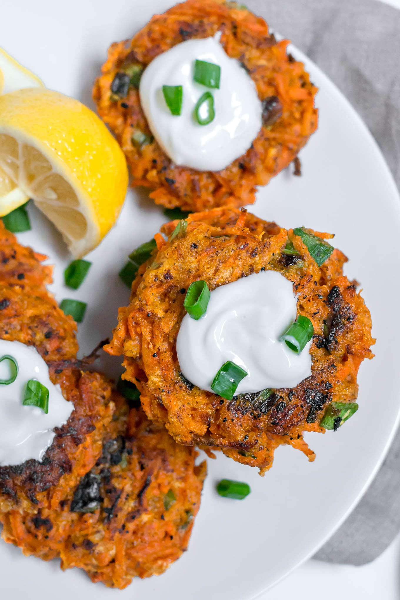 An aerial view of gluten-free carrot fritters on a white plate topped with greek yogurt, and green onions with a lemon wedge.