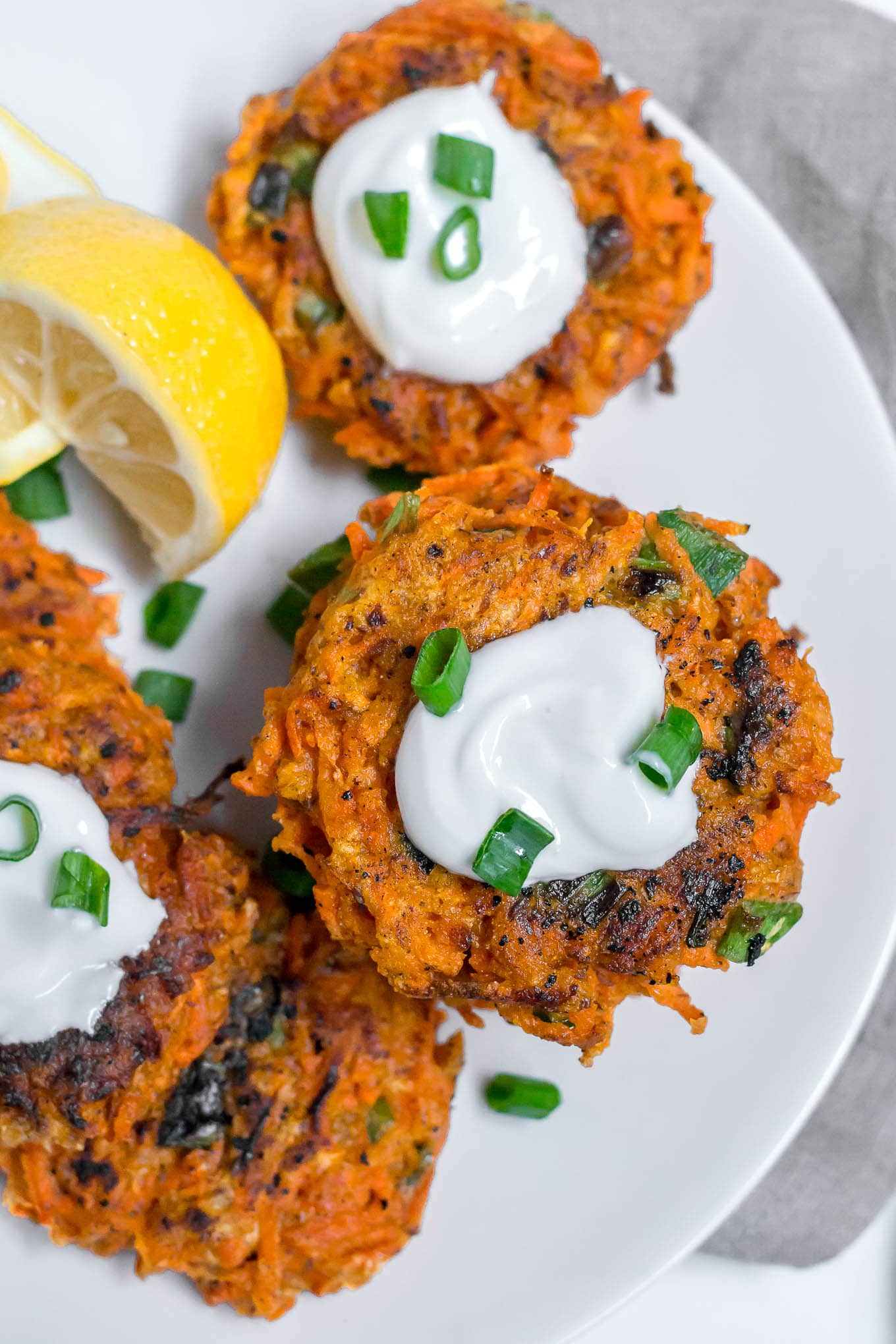 An aerial view of gluten-free carrot fritters on a white plate topped with greek yogurt, and green onions with a lemon wedge. 