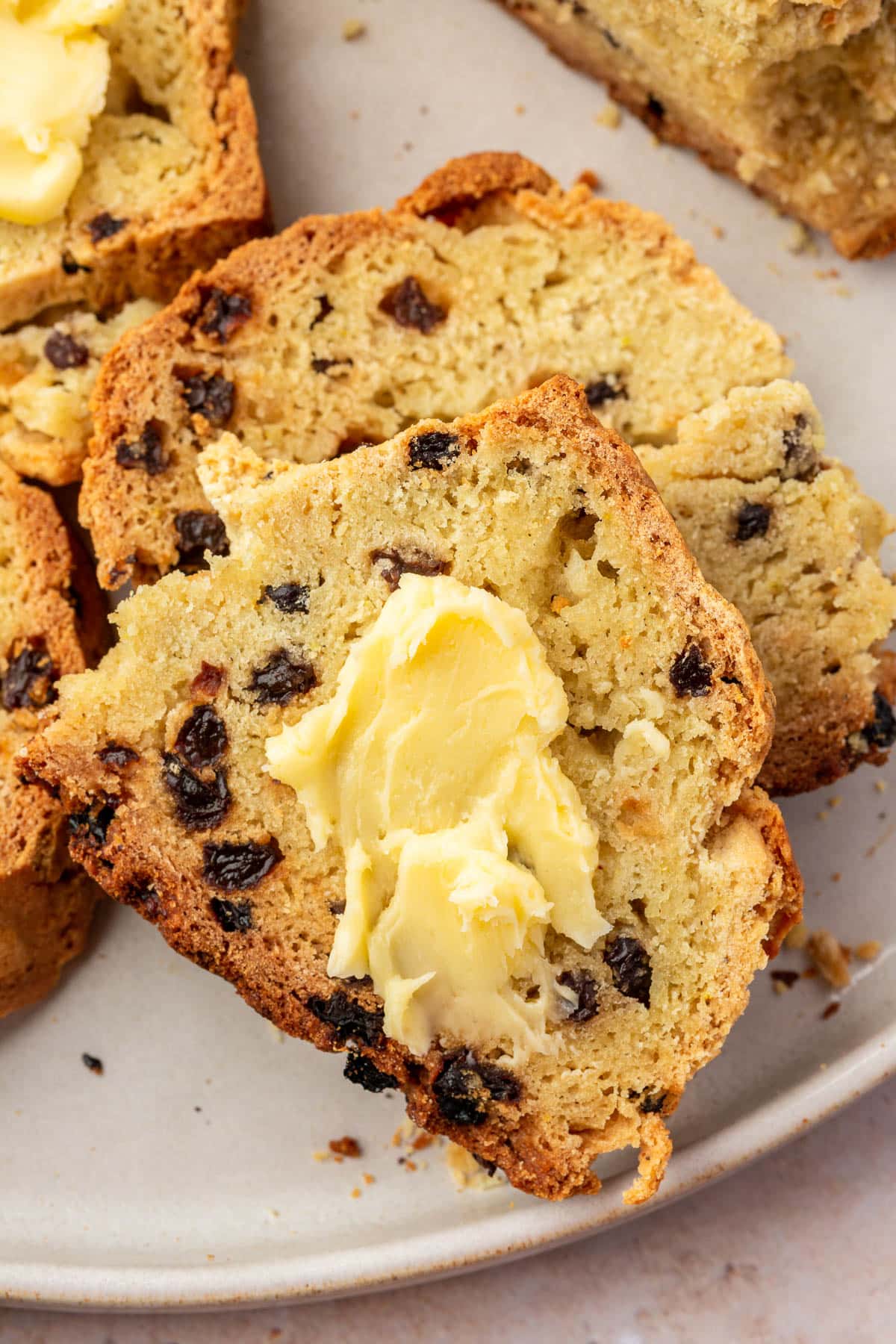 A photo of two slices of gluten-free Irish soda bread with currants topped with a slather of Irish butter with a loaf of Irish soda bread in the background.