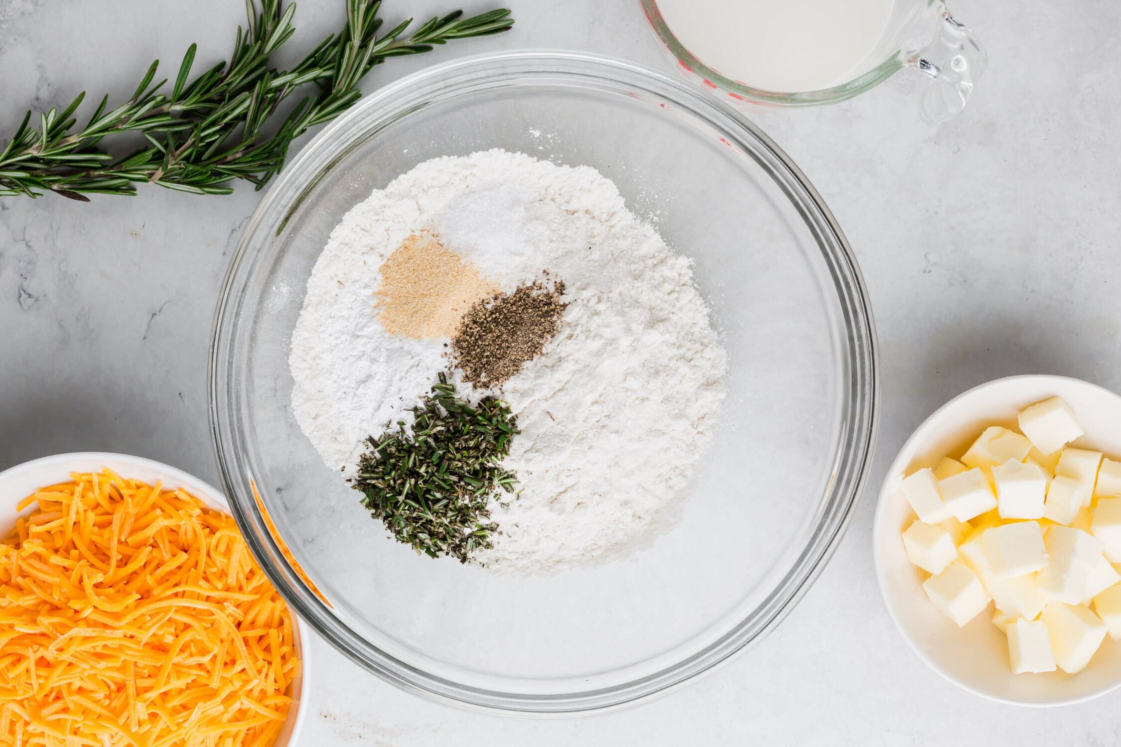 An overhead photo of a bowl of gluten-free flour with salt, baking soda, baking powder, sugar, garlic powder, and rosemary to make gluten-free cheddar biscuits.