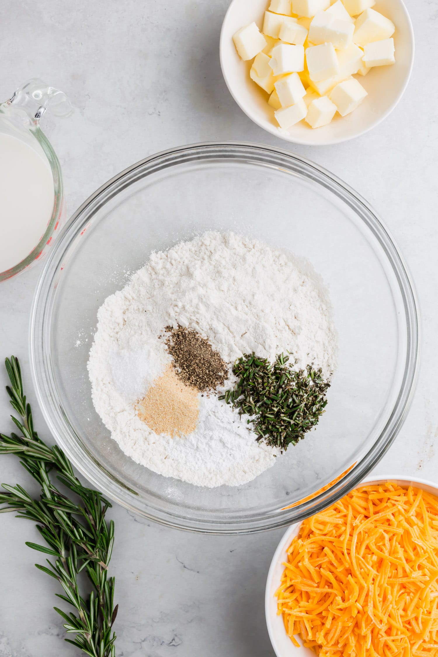 An overhead photo of a bowl of gluten-free flour with salt, baking soda, baking powder, sugar, garlic powder, and rosemary to make gluten-free cheddar biscuits. 