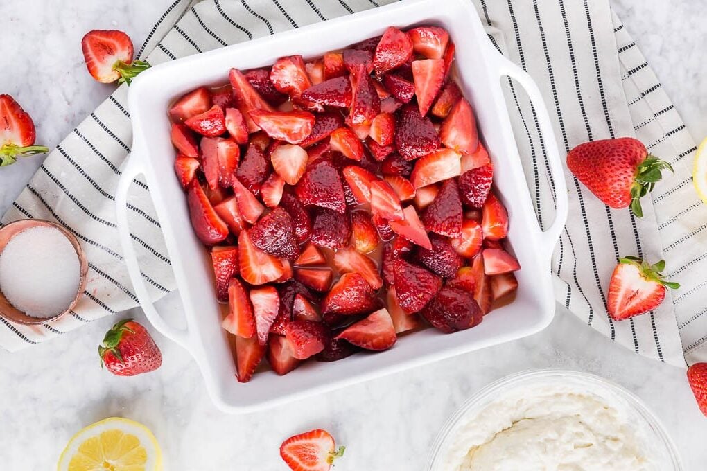A white baking dish filled with chopped strawberries for a gluten-free strawberry cobbler sits on a striped towel, surrounded by whole strawberries, a bowl of sugar, a lemon slice, and whipped topping on a marble surface.