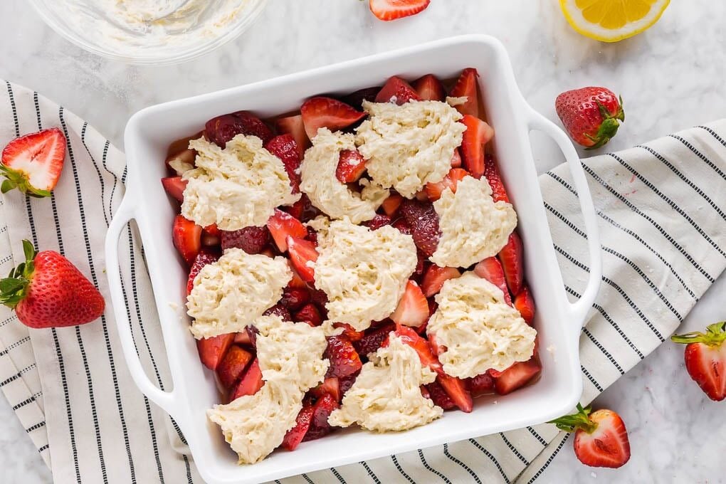 A white baking dish filled with sliced strawberries, topped with dollops of biscuit dough, sits on a striped cloth. Fresh strawberries and a lemon wedge are scattered around this inviting gluten-free strawberry cobbler.
